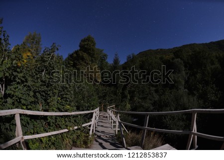 wooden old bridge in forest over treetops and wild river in dark night with stars and milky way in sky