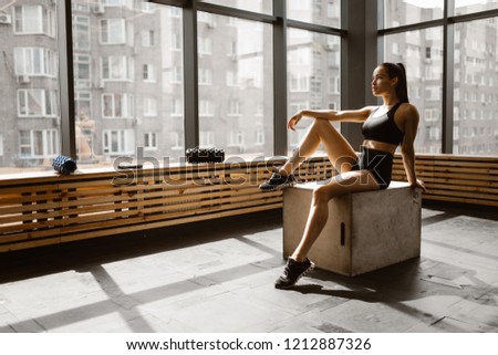 Charming slander girl dressed in black sports top and shorts is sitting on a wooden box in the sunlight in front the window in the gym