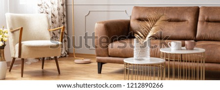 Panoramic view of living room with two small coffee tables, vintage armchair and brown leather sofa, real photo
