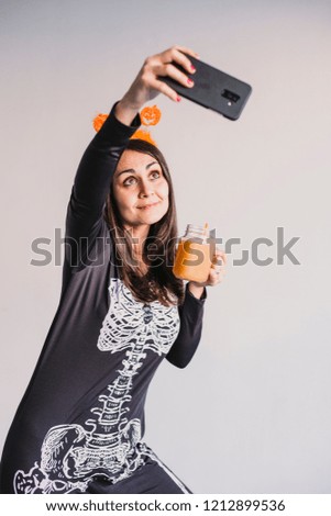 young beautiful woman drinking orange juice and taking a selfie with mobile phone. Wearing a black and white skeleton costume. Halloween concept. Indoors