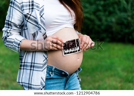 Crop of stylish pregnant woman in jeans and checked shirt holding first photo screen of baby. Brunette mom touching, embracing stomach posing in park. Mother spending time outdoors.