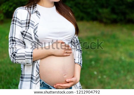 Crop of stylish pregnant woman in jeans and checked shirt holding hands on tummy. Brunette mom touching, embracing stomach posing in park. Mother spending time outdoors.