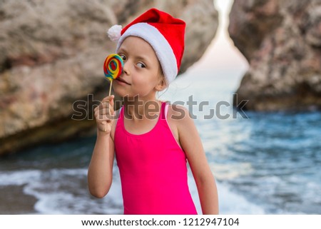 Christmas background: cute child in Santa hat celebrating New Year and Christmas on the beach, free space. Winter holidays in tropical countries. The girl in the Christmas hat and with a lollipop.