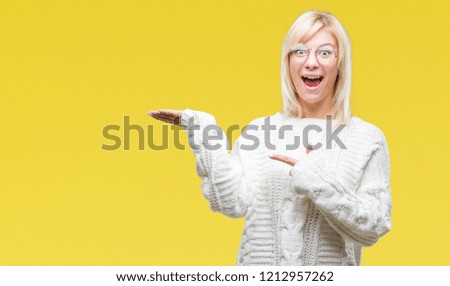 Young beautiful blonde woman wearing winter sweater and glasses over isolated background amazed and smiling to the camera while presenting with hand and pointing with finger.