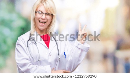 Young beautiful blonde doctor woman wearing medical uniform over isolated background smiling with happy face winking at the camera doing victory sign. Number two.