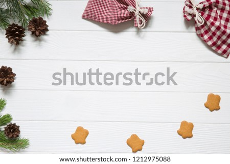 Christmas holidays background, copy space. Fir and pine tree branches with pinecones, christmas chequered red and white bags, ginger cookies on white wooden background, top view. 