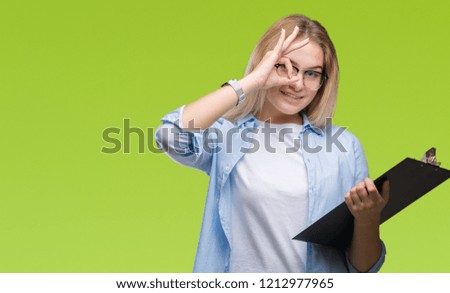 Young caucasian business woman holding clipboard over isolated background with happy face smiling doing ok sign with hand on eye looking through fingers