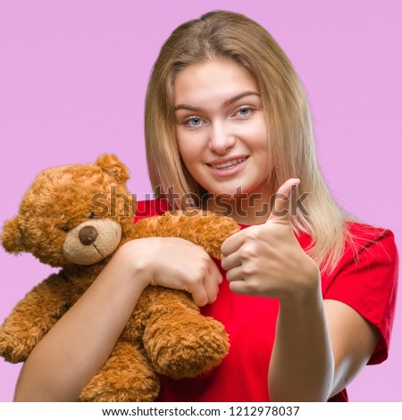 Young caucasian woman holding cute teddy bear over isolated background happy with big smile doing ok sign, thumb up with fingers, excellent sign