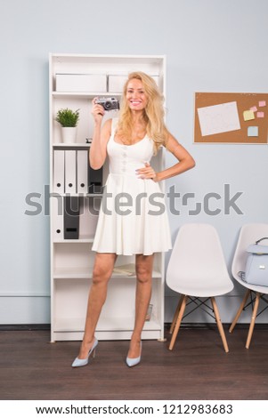 Full length portrait of a smiling pretty girl in dress taking photo on a retro camera in coworking space background
