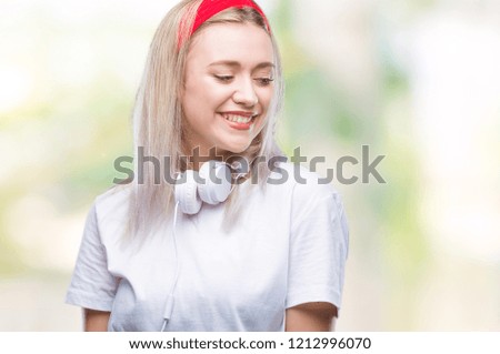 Young blonde woman wearing headphones listening to music over isolated background looking away to side with smile on face, natural expression. Laughing confident.