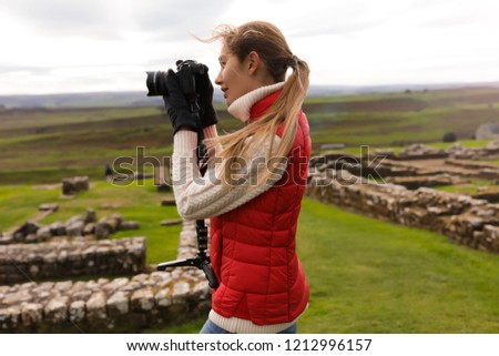 Beautiful woman photographer standing near the ancient ruins. Wearing  the red waistcoat and gloves. Trying to take good pictures of the nature. Green field around her. Stones on the background. 