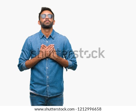 Adult hispanic man wearing sunglasses over isolated background smiling with hands on chest with closed eyes and grateful gesture on face. Health concept.