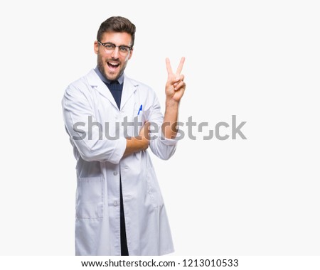 Young handsome man wearing doctor, scientis coat over isolated background smiling with happy face winking at the camera doing victory sign. Number two.