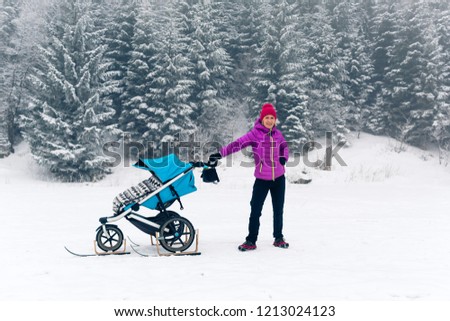 Active mother with baby stroller enjoying motherhood in winter forest, mountains landscape. Jogging or power walking woman with sledge pram in woods. Beautiful winter snowy inspirational mountains.