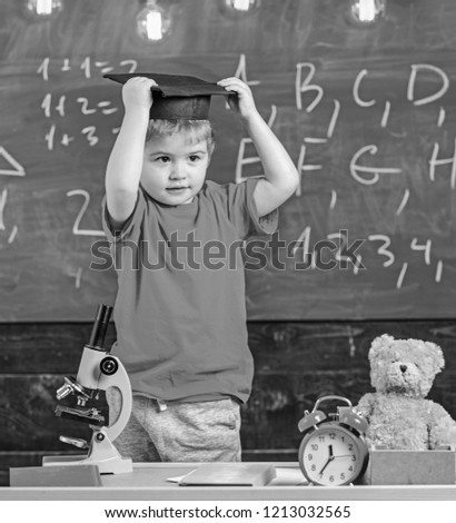 Kid boy in graduate cap ready to go to school, chalkboard on background. Child, pupil on smiling face near microscope. First former interested in studying, education. Kindergarten graduation concept.