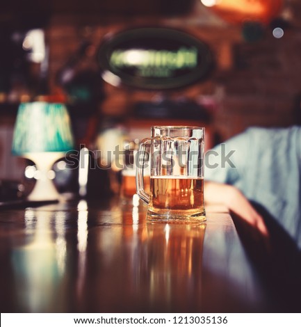 Glass of light beer on wooden bar counter. Man sits behind glass of light beer. Service and alcohol concept. Glass of draft beer on blurred bar background.