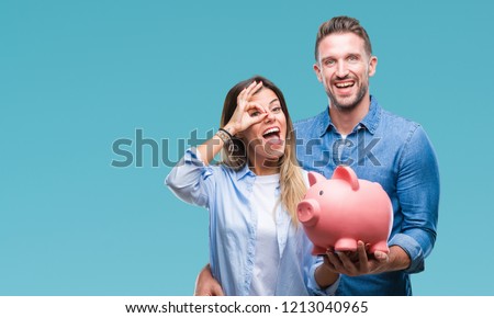 Young couple in love holding piggy bank over isolated background with happy face smiling doing ok sign with hand on eye looking through fingers