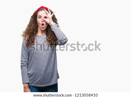 Beautiful brunette curly hair young girl wearing stripes sweater over isolated background doing ok gesture shocked with surprised face, eye looking through fingers. Unbelieving expression.