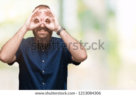 Young caucasian hipster man over isolated background doing ok gesture like binoculars sticking tongue out, eyes looking through fingers. Crazy expression.