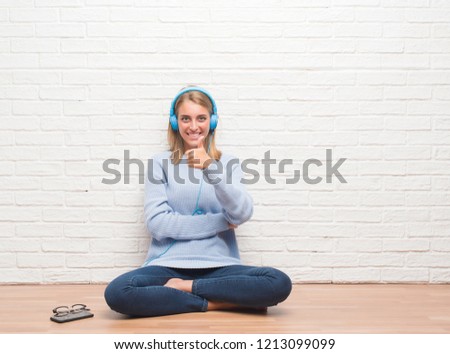 Beautiful young woman sitting on the floor listening music wearing headphones at home happy with big smile doing ok sign, thumb up with fingers, excellent sign