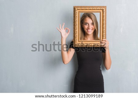 Beautiful young woman over grunge grey wall holding vintage frame doing ok sign with fingers, excellent symbol