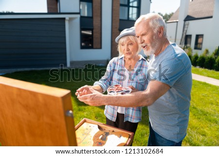 Taking brush. Bearded elderly man taking painting brush while helping his appealing beaming wife drawing nature