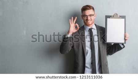 Young redhead business man over grey grunge wall holding clipboard doing ok sign with fingers, excellent symbol