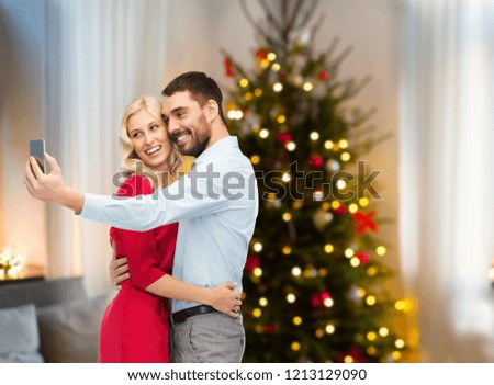 technology and holidays concept - happy couple taking selfie by smartphone over home room with christmas tree background