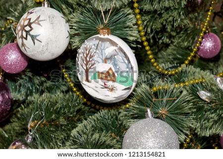 Close up of Christmas tree and unique handmade glass toy with picture of house in the snow and pine cones and lights and ball with snowflake.