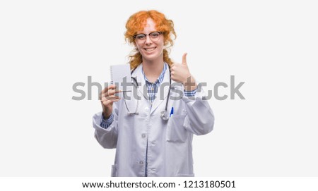 Young redhead doctor woman holding blank notebook happy with big smile doing ok sign, thumb up with fingers, excellent sign
