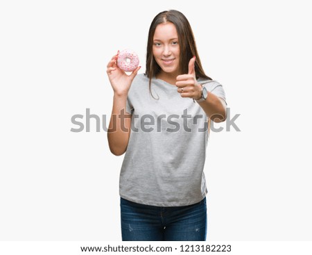 Young caucasian woman eating sweet donut over isolated background happy with big smile doing ok sign, thumb up with fingers, excellent sign