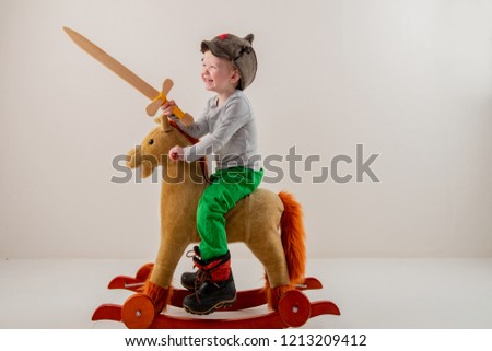 Funny child in old military cap with red star, on toy horse-rocking horse with wooden sword. Boy dreams of battles, victories and adventures. Concept training of spirit, education morale, patriotizm 
