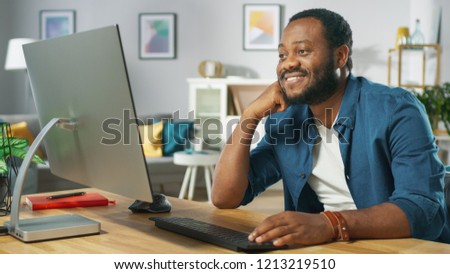 Portrait of the Happy Man Sitting at His Desk, Watching Videos at Home. In the Background Cozy Living Room.