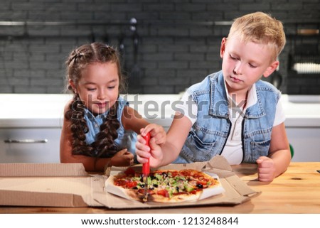 Two children: a boy and a girl in the kitchen cut pizza. Photo in interior.