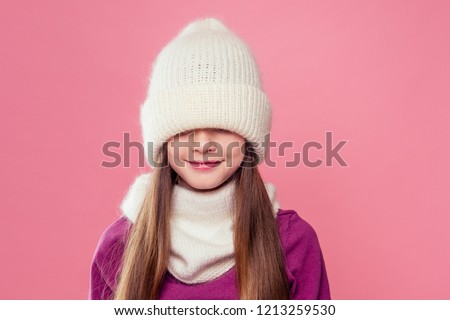 little girl in winter warm white hat and scarf and in pink mittens dreaming Christmas night playing hide and seek in studio on pink background.portrait photography child model hiding eyes under cap