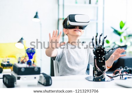 Nice smart enthusiastic boy sitting at the table and wearing VR glasses while experimenting with his robotic devices Royalty-Free Stock Photo #1213293706
