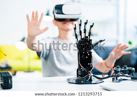 Selective focus of a robotic hand on the table with a little smiling boy testing VR glasses in the background Royalty-Free Stock Photo #1213293715