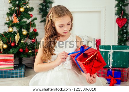 Cute little girl in a nice dress with long curly blondу hair dreaming and holding a box with a gift at home near a Christmas tree with gifts and garlands and a decorated fireplace .new Year morning