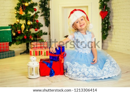 Cute little girl in dress Santa Claus hat curly blondу hair wide open eyes holding a lot of gifts box at home near a Christmas tree with gifts and garlands and a decorated fireplace .new Year morning