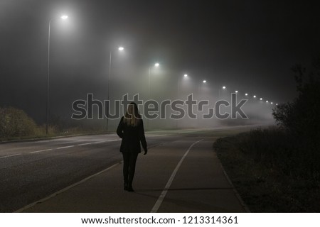 Young woman alone slowly walking under white street lights in night. Dark time. Peaceful atmosphere in mist. Foggy air. Back view. Royalty-Free Stock Photo #1213314361