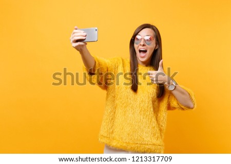 Excited young woman in fur sweater, heart glasses showing thumb up, doing taking selfie shot on mobile phone isolated on bright yellow background. People sincere emotions, lifestyle. Advertising area