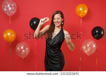 Joyful girl in black dress doing winner gesture holding bitcoin metal coin of golden color future currency on bright red background air balloon. Happy New Year, birthday mockup holiday party concept