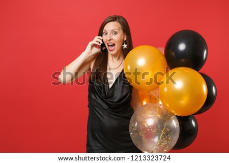 Excited young girl in little black dress holding air balloons talking on mobile phone conducting pleasant conversation isolated on red background. Happy New Year birthday mockup holiday party concept
