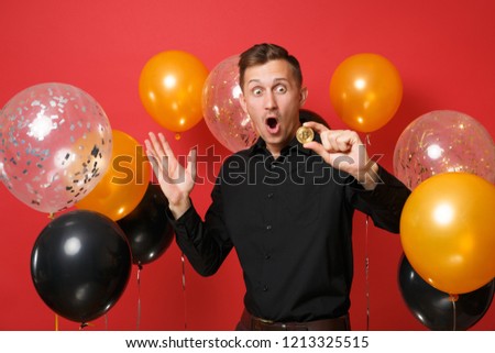 Shocked young man in classic shirt spreading hands, holding bitcoin, metal coin of golden color, future currency on red background air balloons. Happy New Year, birthday mockup holiday party concept