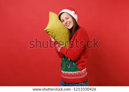 Cheerful young Santa girl in Christmas hat holding pillow, pretending asleep blinking isolated on bright red wall background. Happy New Year 2019 celebration holiday party concept. Mock up copy space
