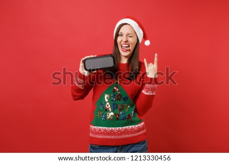 Santa girl showing horns gesture, depicting heavy metal rock sign, holding portable wireless bluetooth music speaker isolated on red background. Happy New Year 2019 celebration holiday party concept
