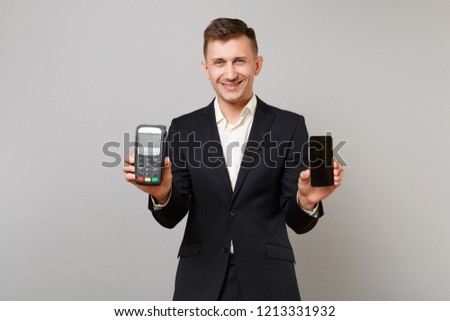Smiling business man holding wireless modern bank payment terminal to process, acquire credit card payments, mobile phone isolated on grey wall background. Achievement career wealth business concept
