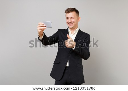 Joyful young business man in classic black suit showing thumb up, doing taking selfie shot on mobile phone isolated on grey background. Achievement career wealth business concept. Mock up copy space