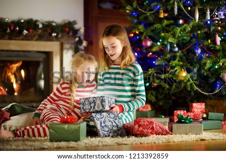 Happy little sisters wearing Christmas pajamas opening gift boxes by a fireplace in a cozy dark living room on Christmas eve. Celebrating Xmas at home.