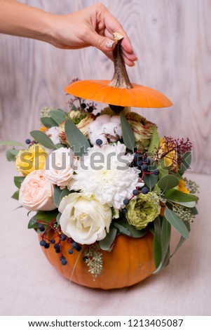 Autumn floral bouquet in a pumpkin vase on a light background, a mixture of flowers, pionic rose, eucalyptus, chrysanthemum. Female hand holding a pumpkin hat